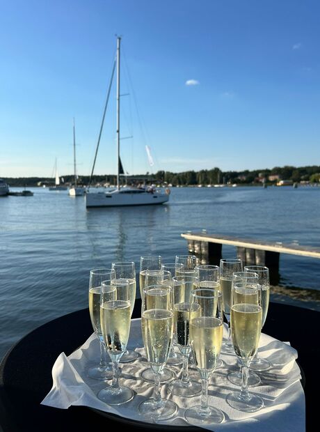 Tablett mit Sektgläsern auf einem Tisch an der Marina, im Hintergrund Segelboote und ruhiges Wasser