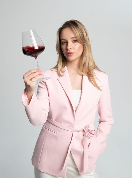 Kobieta w różowej marynarce trzyma kieliszek czerwonego wina, gotowa na degustację w hotelowym barze