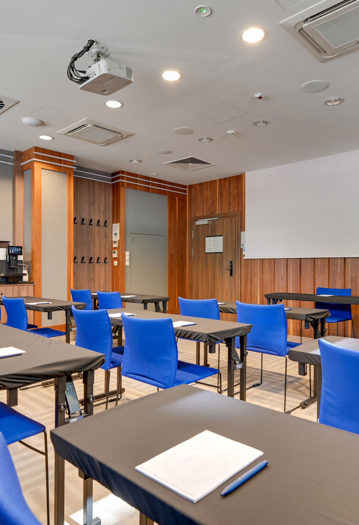 Konferenzraum mit blauen Stühlen, Reihen-Tischen, Beamer und Leinwand, Holzverkleidung und Kaffee-Servicestation.