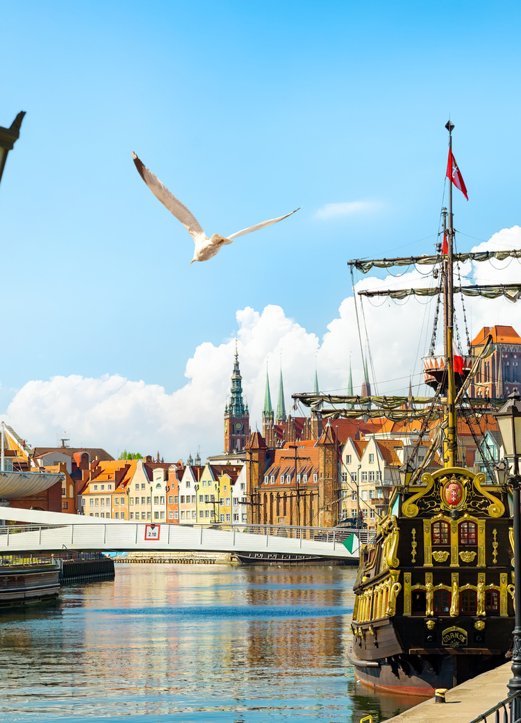 Historic waterfront promenade with a docked ornate tall ship, colorful gabled buildings, church towers and sunny river views.