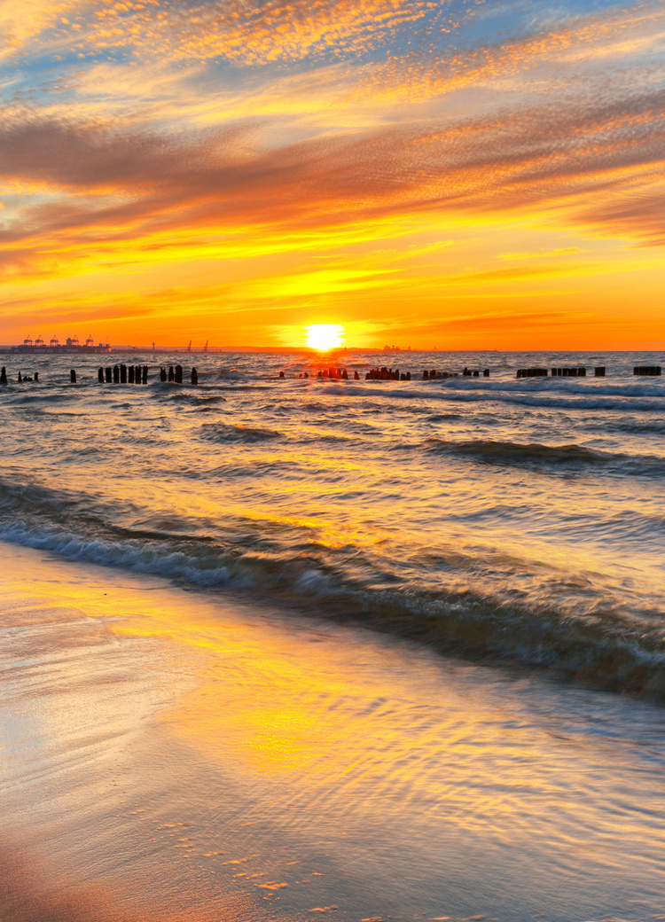 Zachód słońca nad piaszczystą plażą, łagodne fale i drewniane falochrony, złote niebo odbija się na mokrym piasku