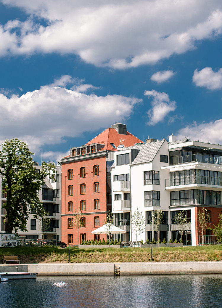 Riverside boutique hotel with modern white and red-brick façade, balconies, outdoor terrace and a moored yacht.