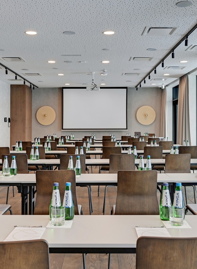 Moderne hotellkonferanserom i klasseromsoppsett med lerret/projektor, rekker av bord og stoler, vannflasker, notatblokker og store vinduer