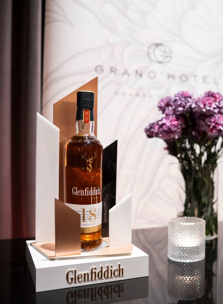 Butelka whisky Glenfiddich na hotelowym barze, kryształowe kieliszki, świeca i fioletowe goździki, elegancki salon