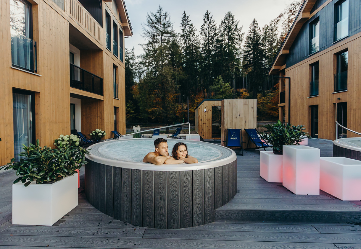 Para w zewnętrznym jacuzzi na drewnianym tarasie między nowoczesnymi drewnianymi budynkami, widok na las i podświetlane donice.