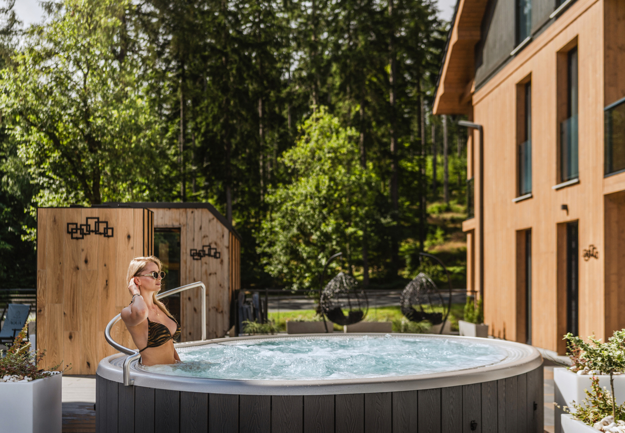 Woman relaxing in an outdoor bubbling hot tub on a hotel's wooden terrace, with forest view and hanging lounge chairs.