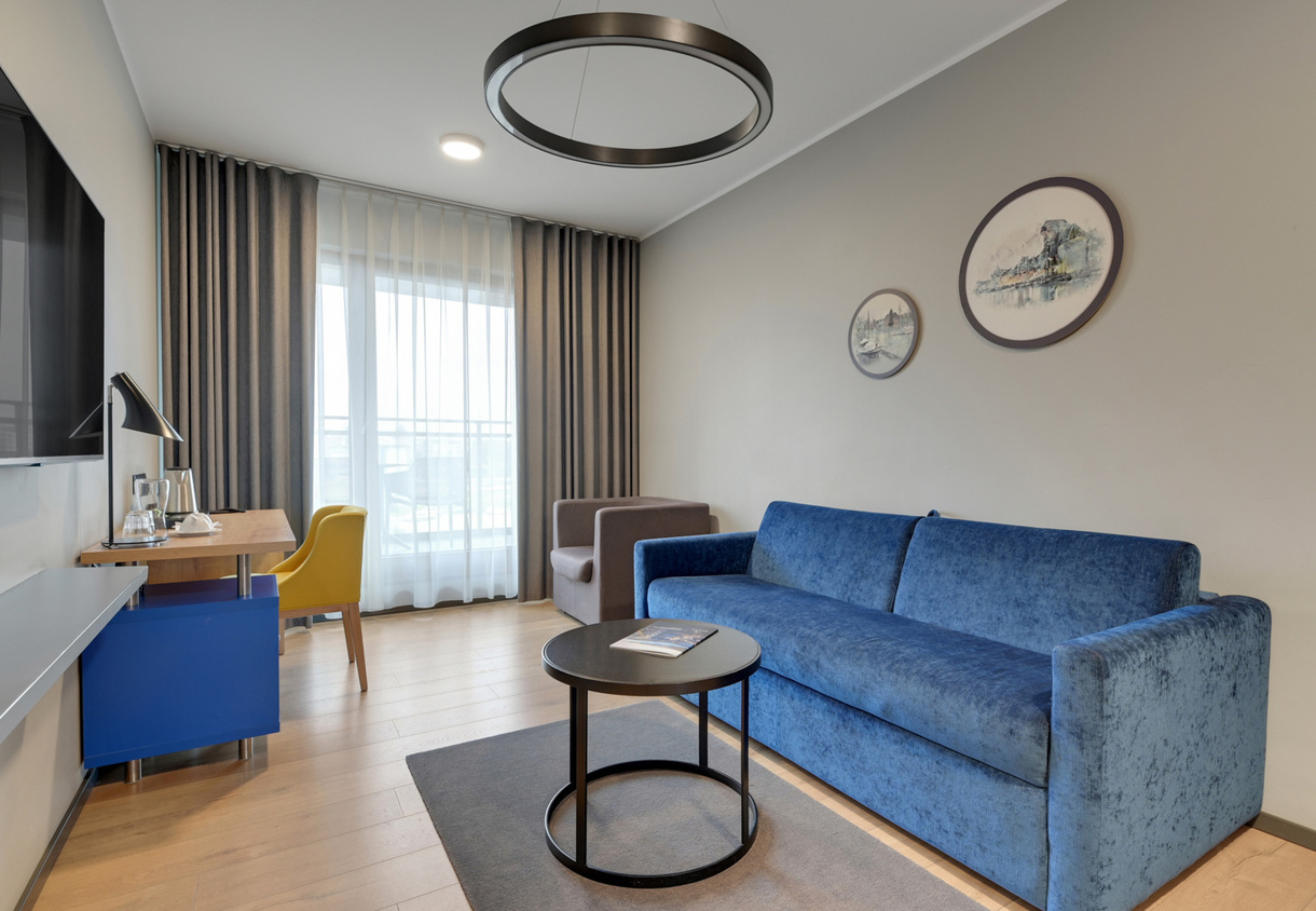 Modern hotel lounge with blue velvet sofa, round coffee table, desk with yellow chair, balcony window and sheer drapes.