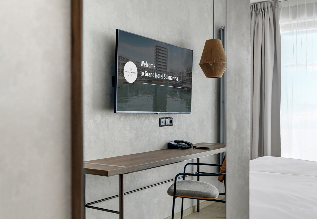 Modern hotel workspace with wall TV showing welcome screen, wooden desk with phone, chair, pendant lamp, bed and curtains.