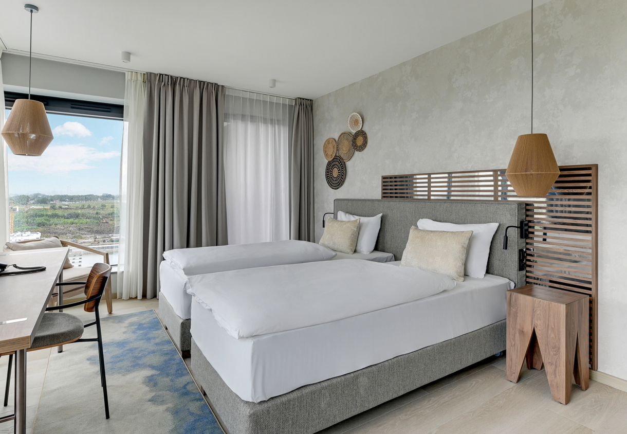 Modern twin hotel room with upholstered headboard, woven pendant lights, wooden side table, desk and large window view.