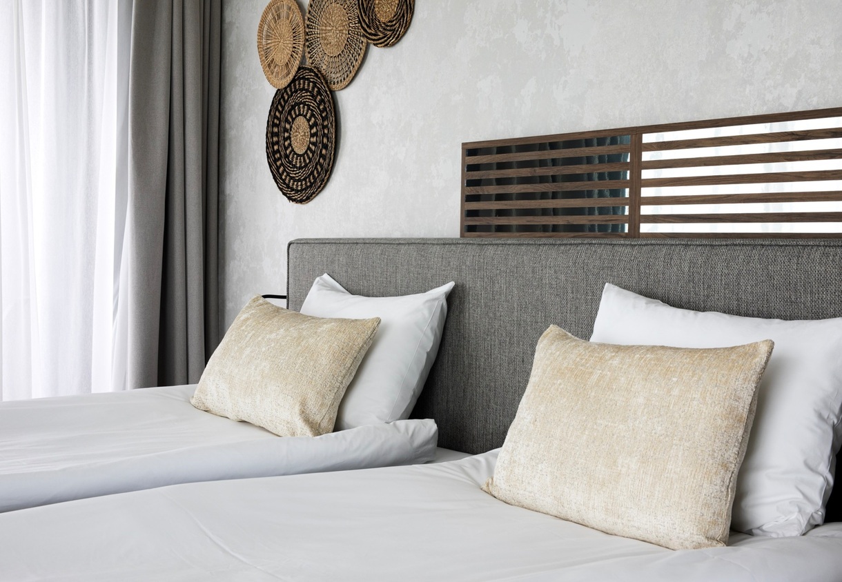 Twin beds with white linens and beige cushions, grey upholstered headboard with slatted wood panel, woven wall decor and sheer curtains