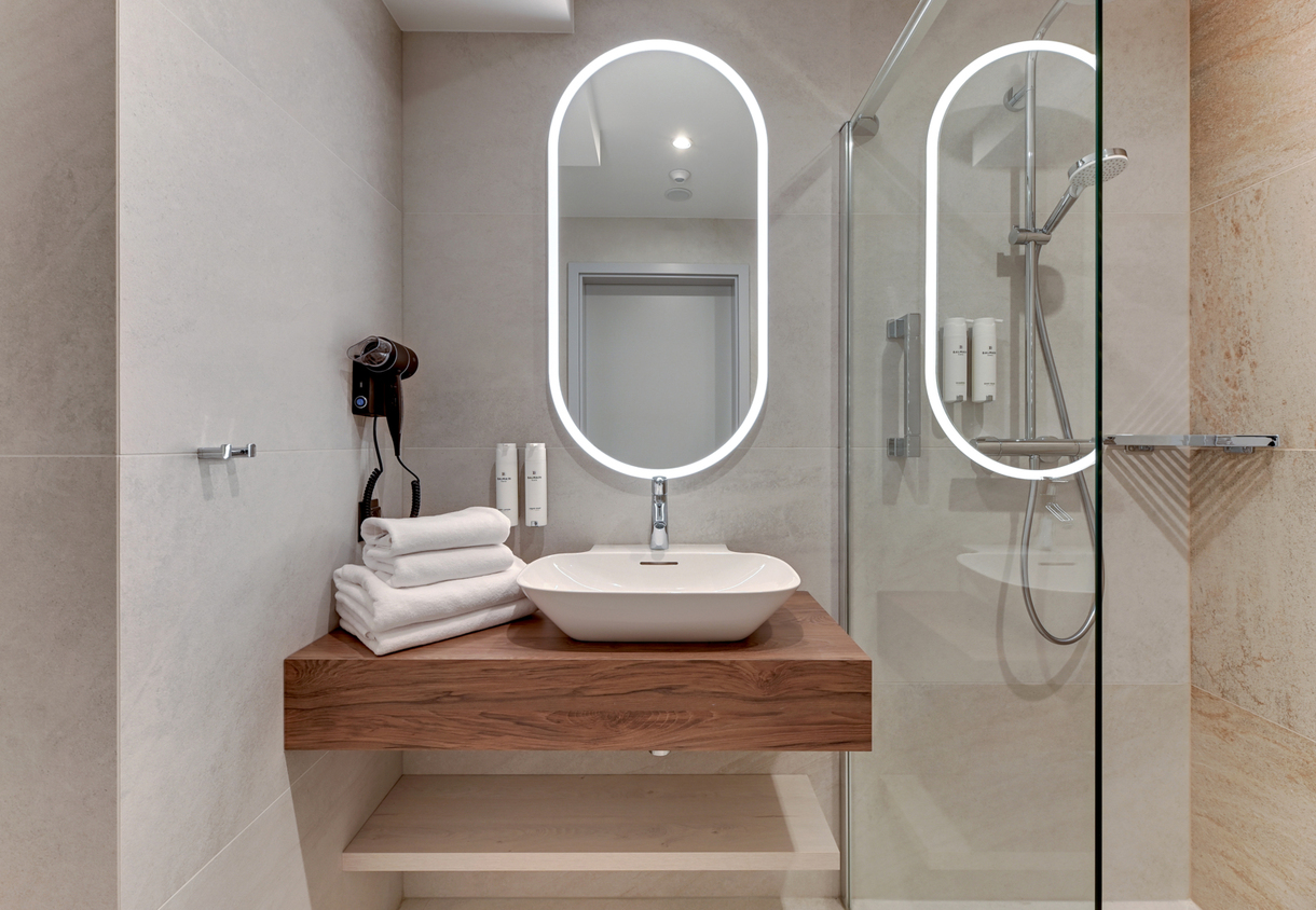 Modern bathroom with backlit oval mirror, vessel sink on wooden vanity, stacked white towels, wall hairdryer and glass shower with rain head.