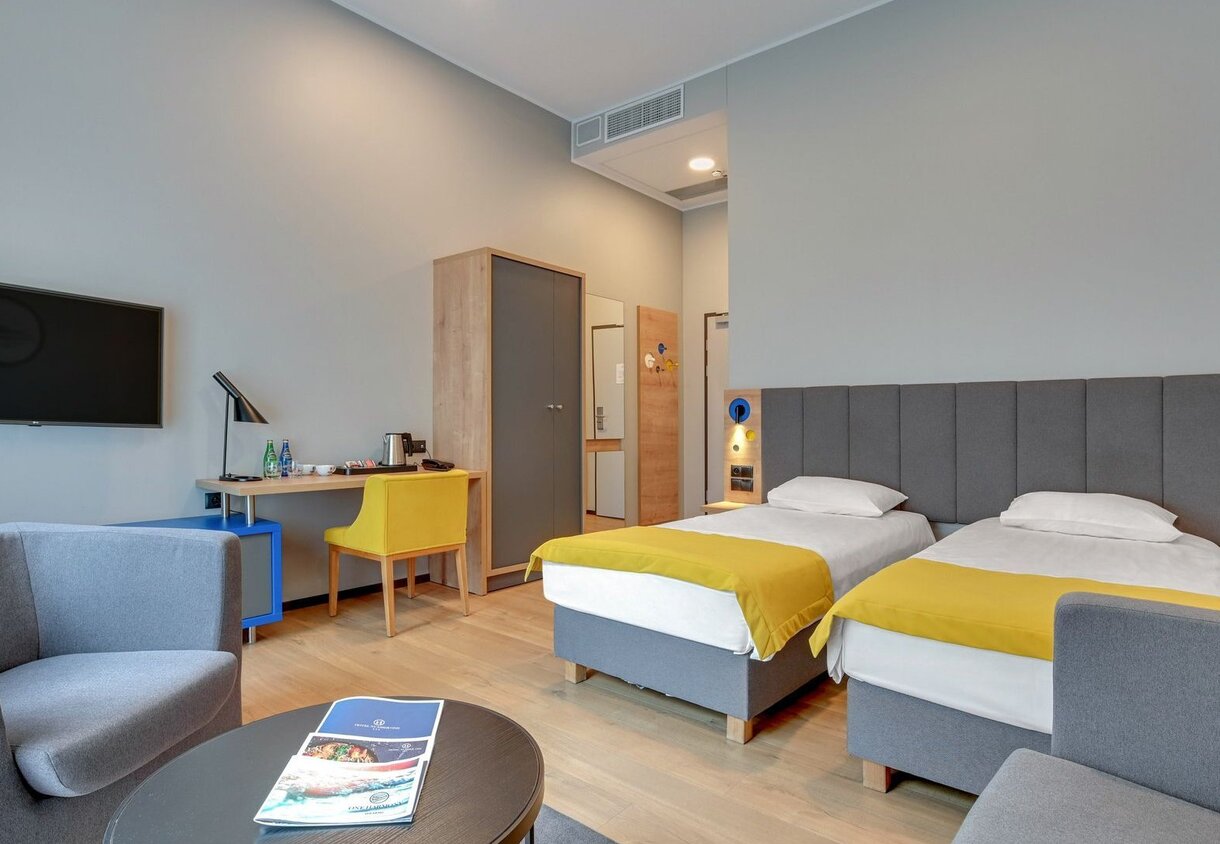 Modern twin hotel room with two single beds, upholstered grey headboard, yellow bed runners, seating area, work desk and wall TV.