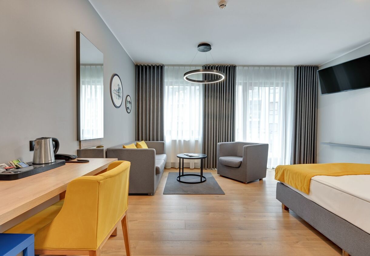 Modern hotel suite with double bed and yellow throw, grey sofa and armchair, ring pendant light, desk with kettle, floor-to-ceiling windows