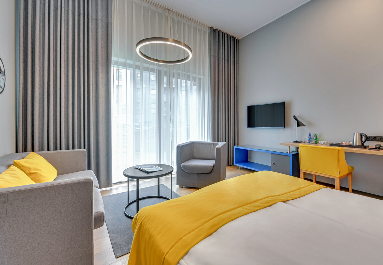 Modern hotel room with yellow bed runner, gray sofa and armchair, floor-to-ceiling curtains, wall TV and desk with kettle.