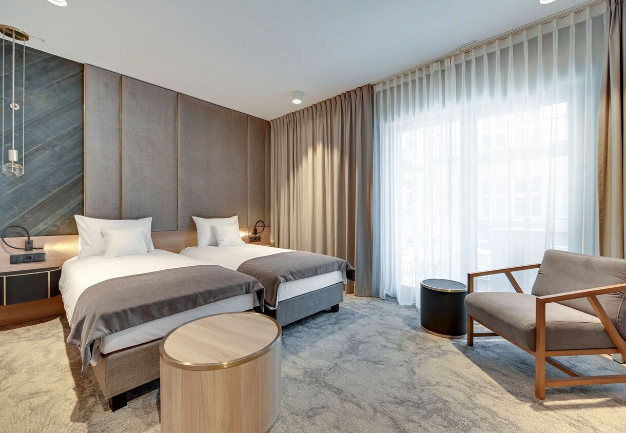 Twin beds with upholstered headboard, bedside reading lights, lounge chair and floor-to-ceiling window with sheer curtains.