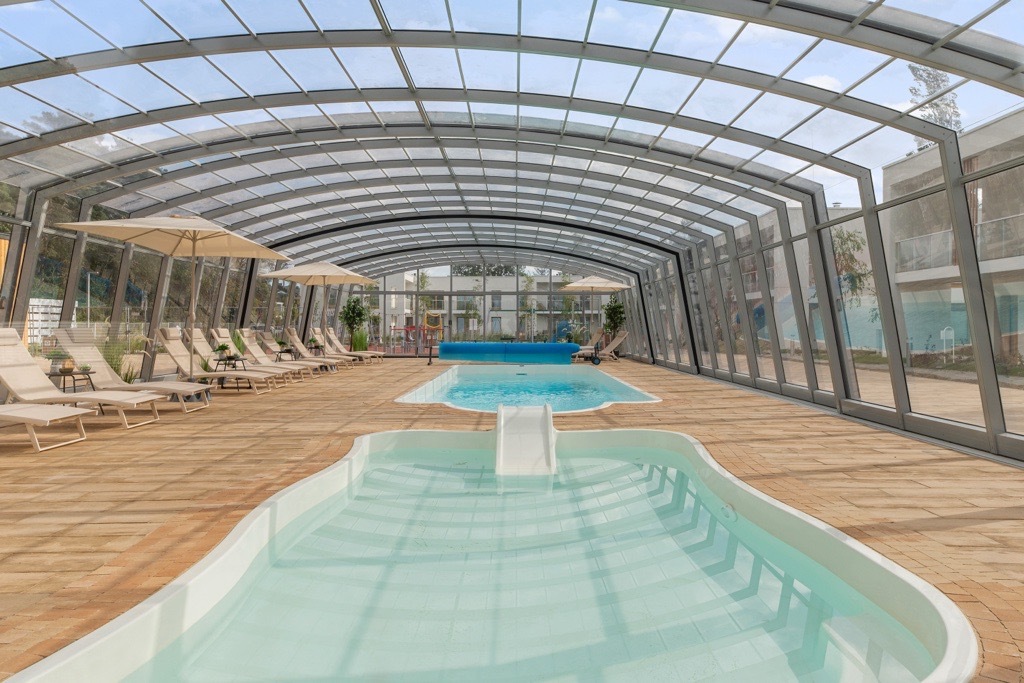 Covered indoor pool complex with glass arched roof, two pools including a shallow kids' pool with slide, lounge chairs and umbrellas.