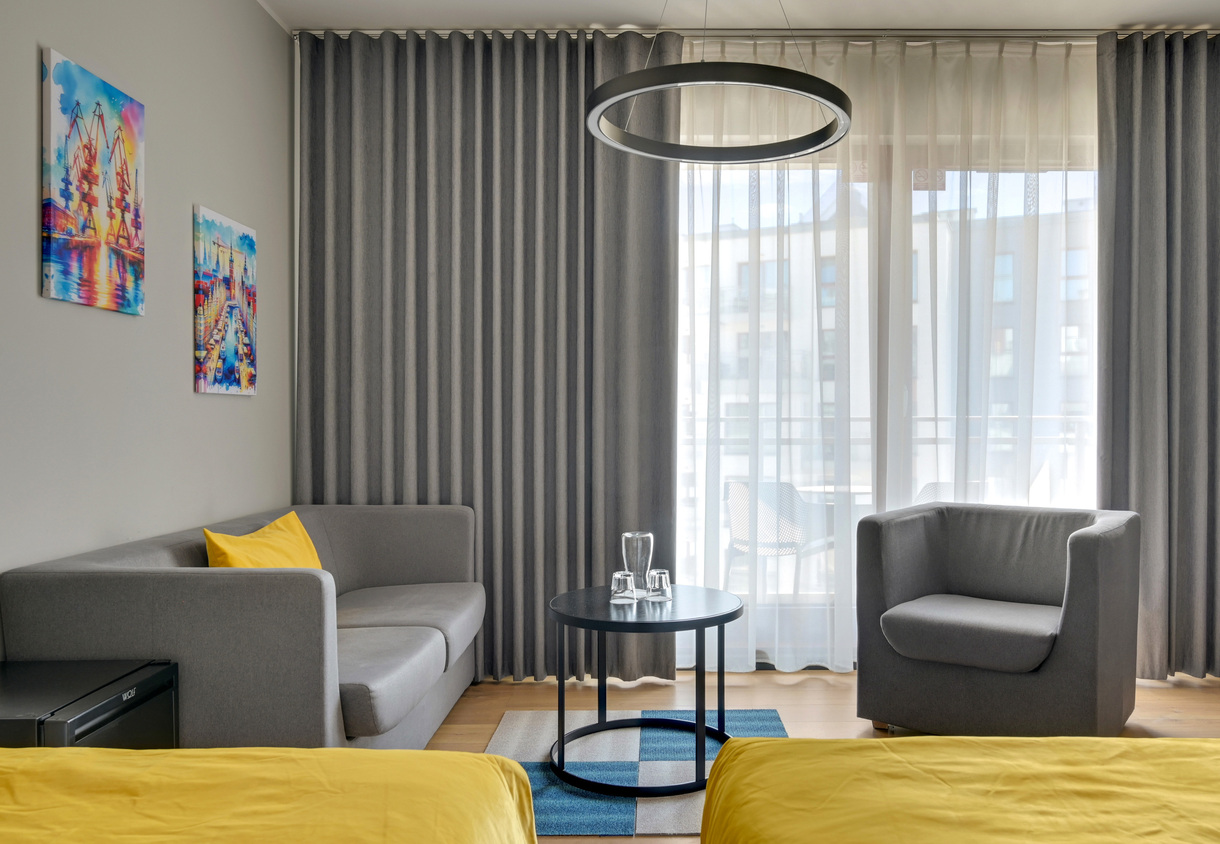 Contemporary hotel lounge with grey sofa and armchair, yellow bedding accents, round black coffee table with glassware, sheer curtains and balcony view