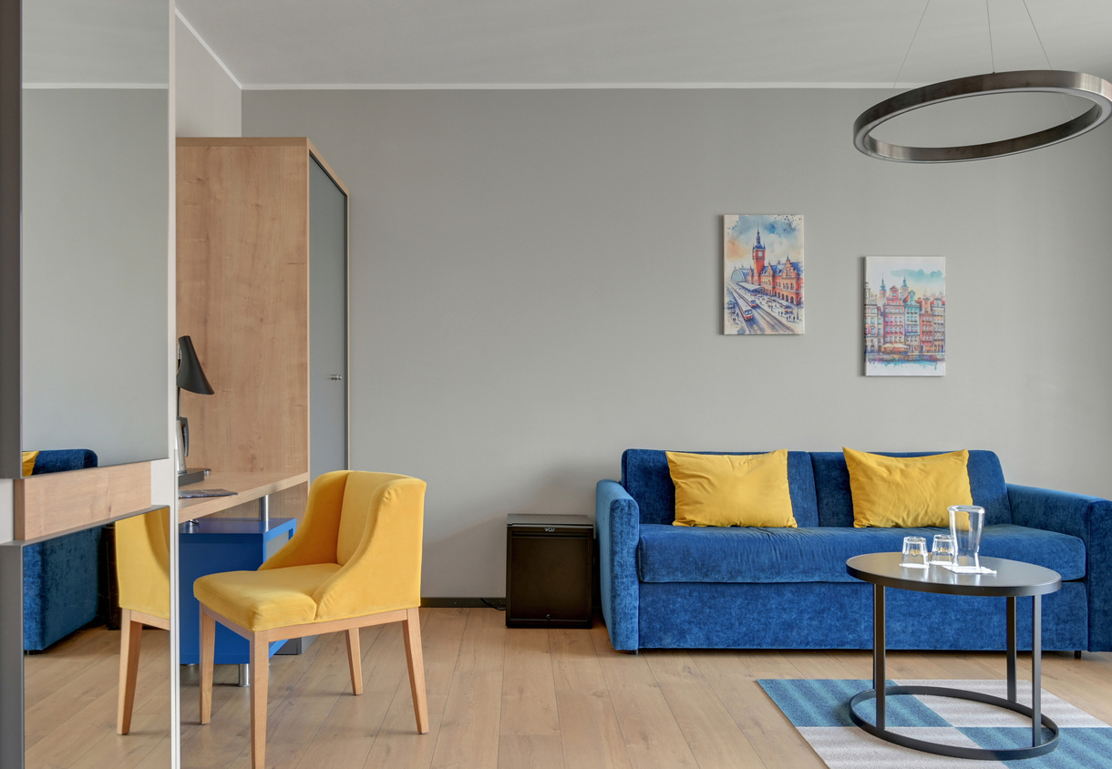 Modern hotel lounge with blue velvet sofa and yellow cushions, yellow armchair, round pendant light and a coffee table with glassware.