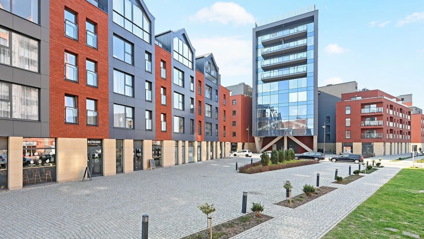 Modern hotel complex with red-brick and glass facades, cobbled courtyard, ground-floor café and shops, landscaped plaza and parking.