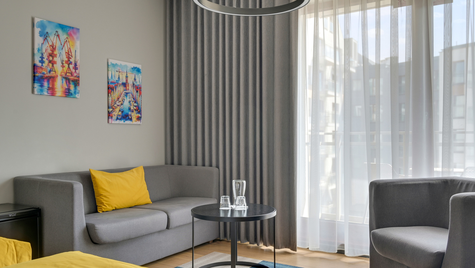 Modern hotel suite with grey sofa, yellow cushions, round black coffee table with glasses, sheer floor-to-ceiling curtains and colorful art.