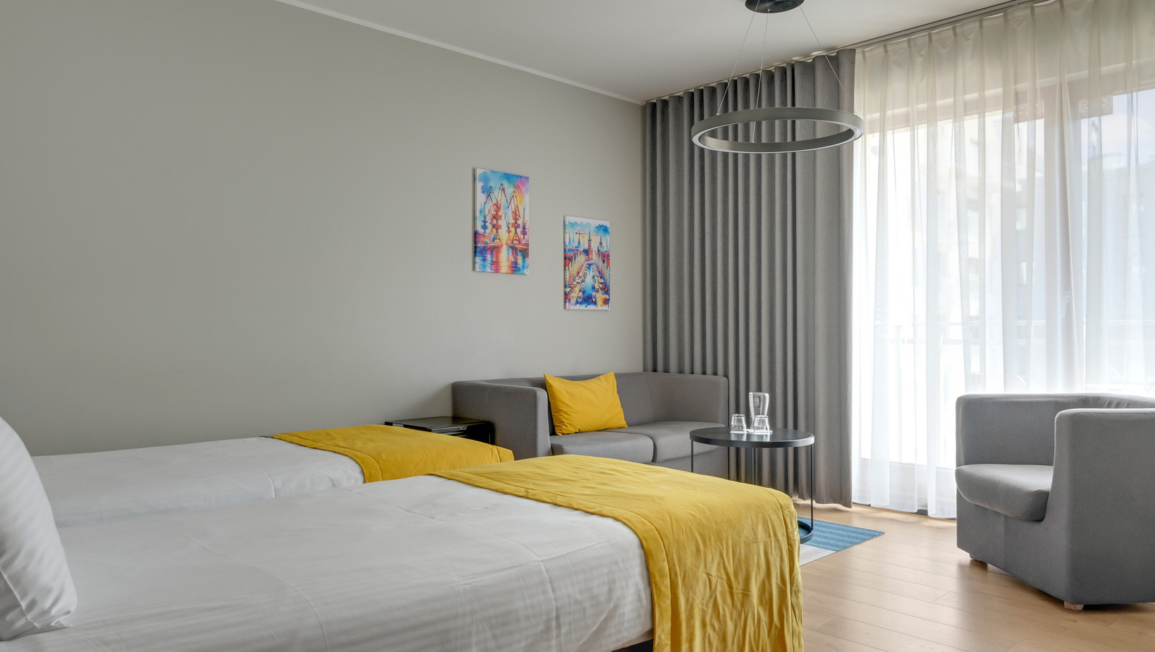 Twin hotel room with white beds and yellow throws, gray sofa with yellow cushion, circular pendant light and sheer curtains.
