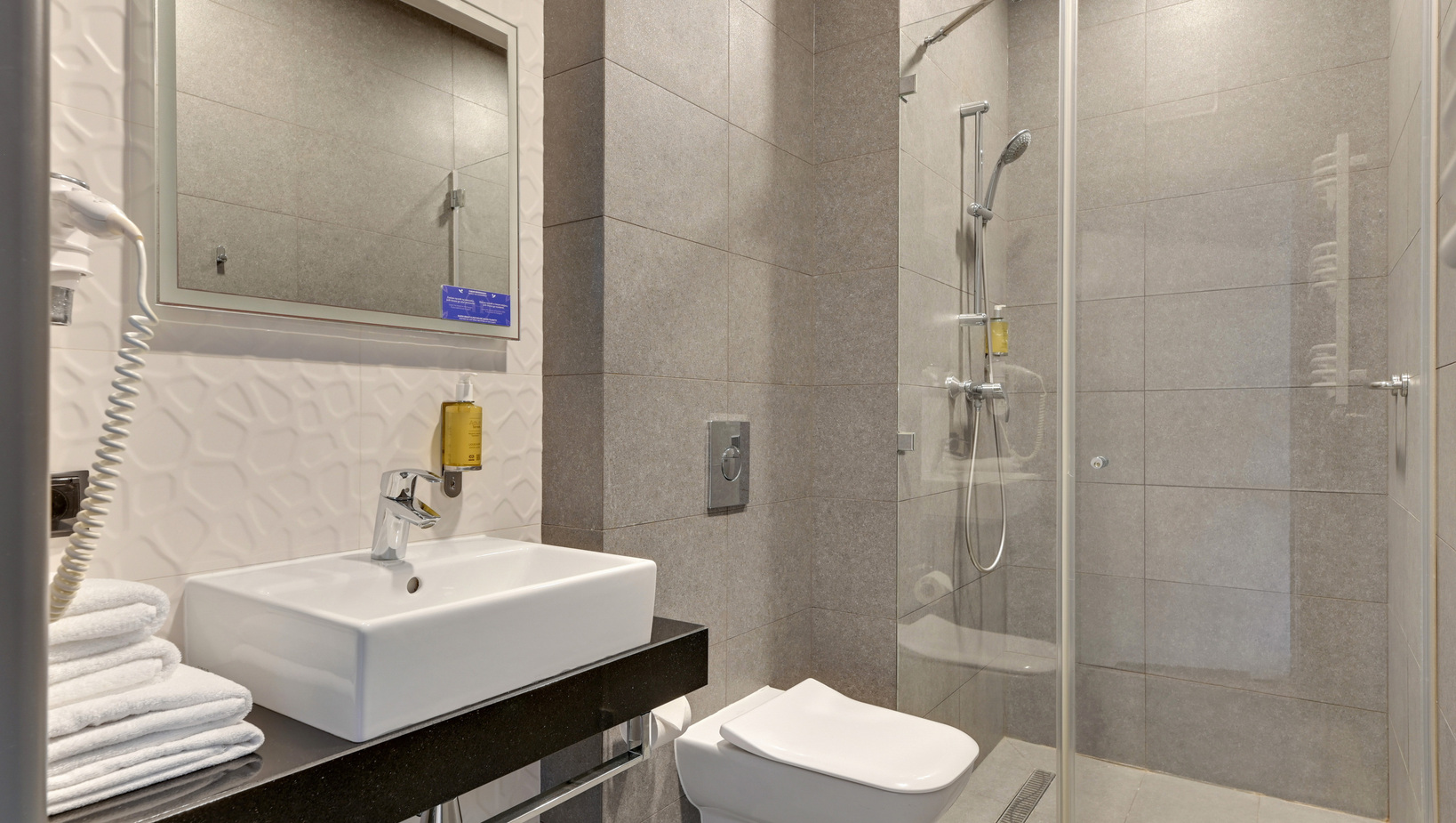 Modern bathroom with glass shower, gray tile walls, wall-mounted toilet, basin on black counter, stacked towels, wall hairdryer and soap.