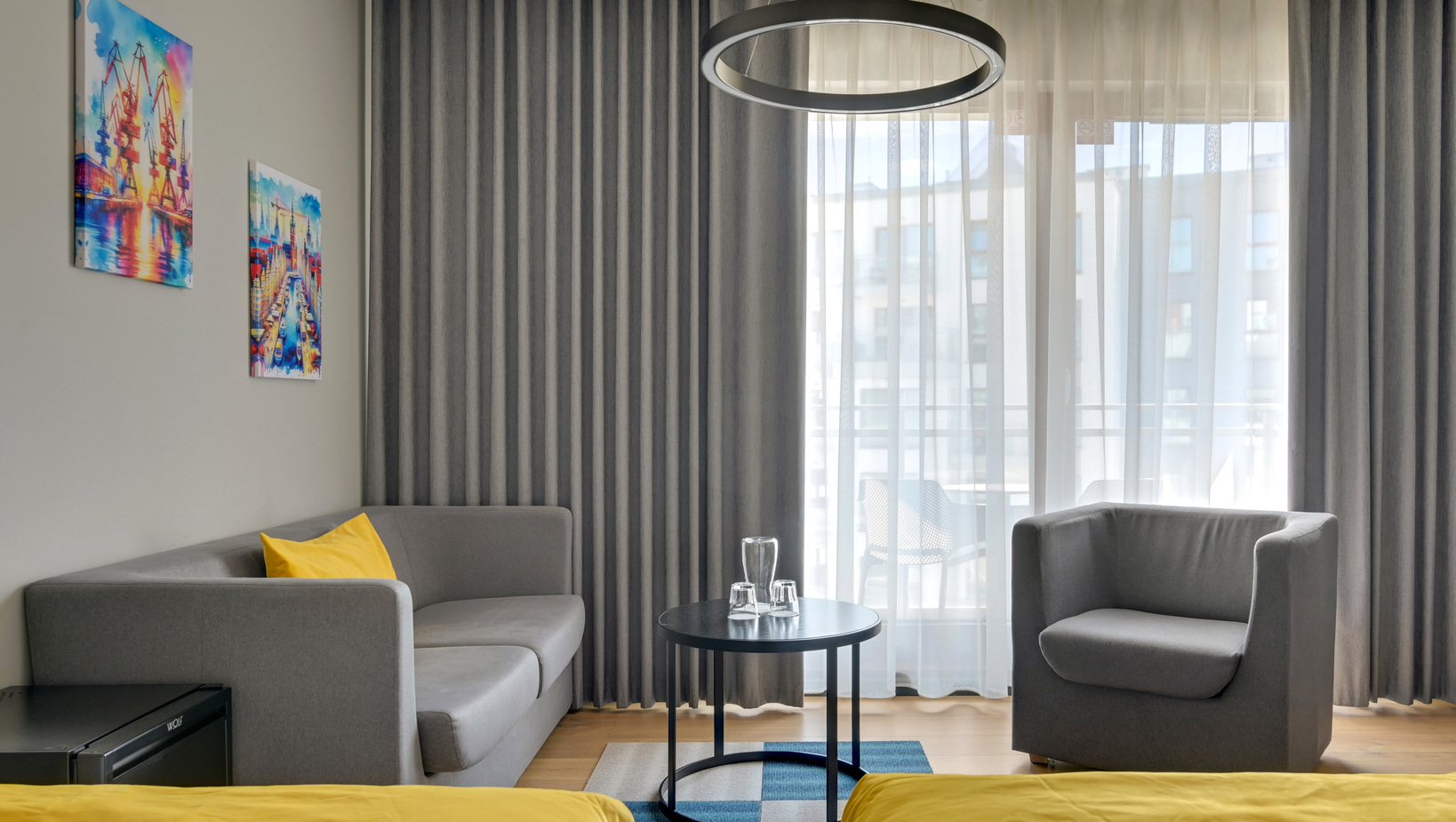 Contemporary hotel lounge with grey sofa and armchair, yellow bedding accents, round black coffee table with glassware, sheer curtains and balcony view