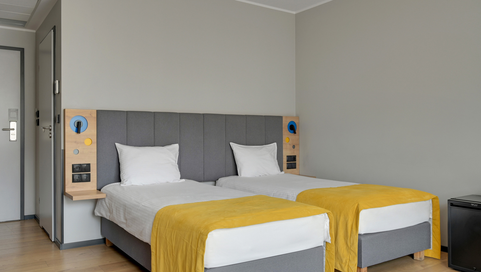 Two twin beds, gray upholstered headboard, white linens and yellow runners, bedside panels with reading lights and outlets.