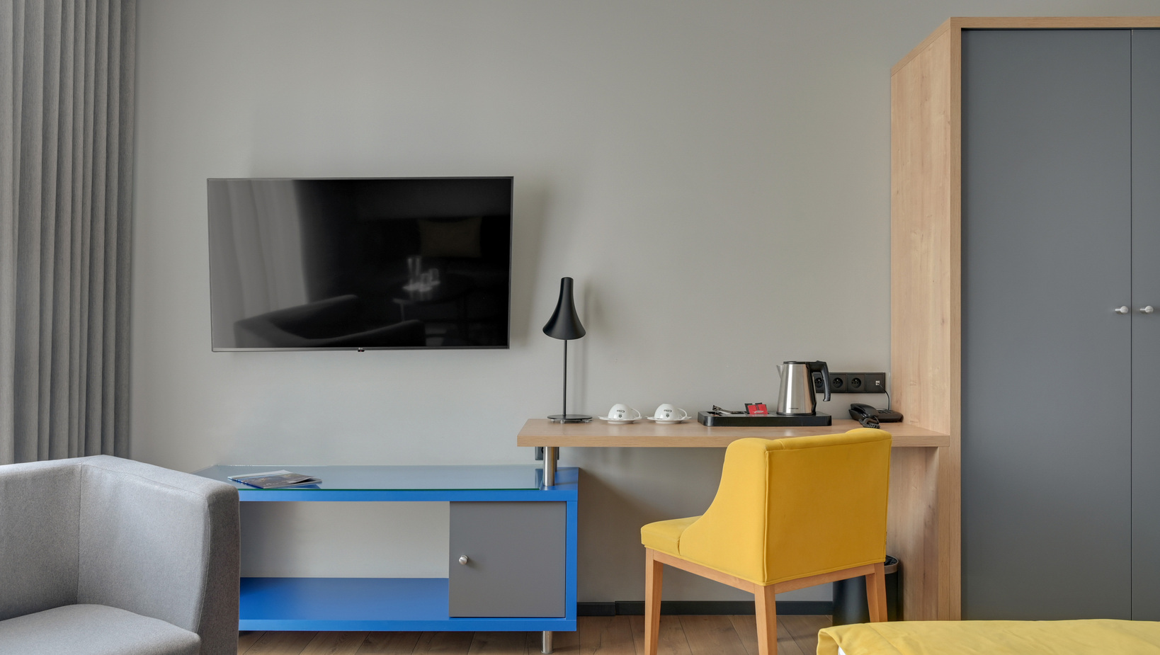 Wall-mounted flat-screen TV above a work desk with electric kettle, cups on a tray, black floor lamp and yellow chair