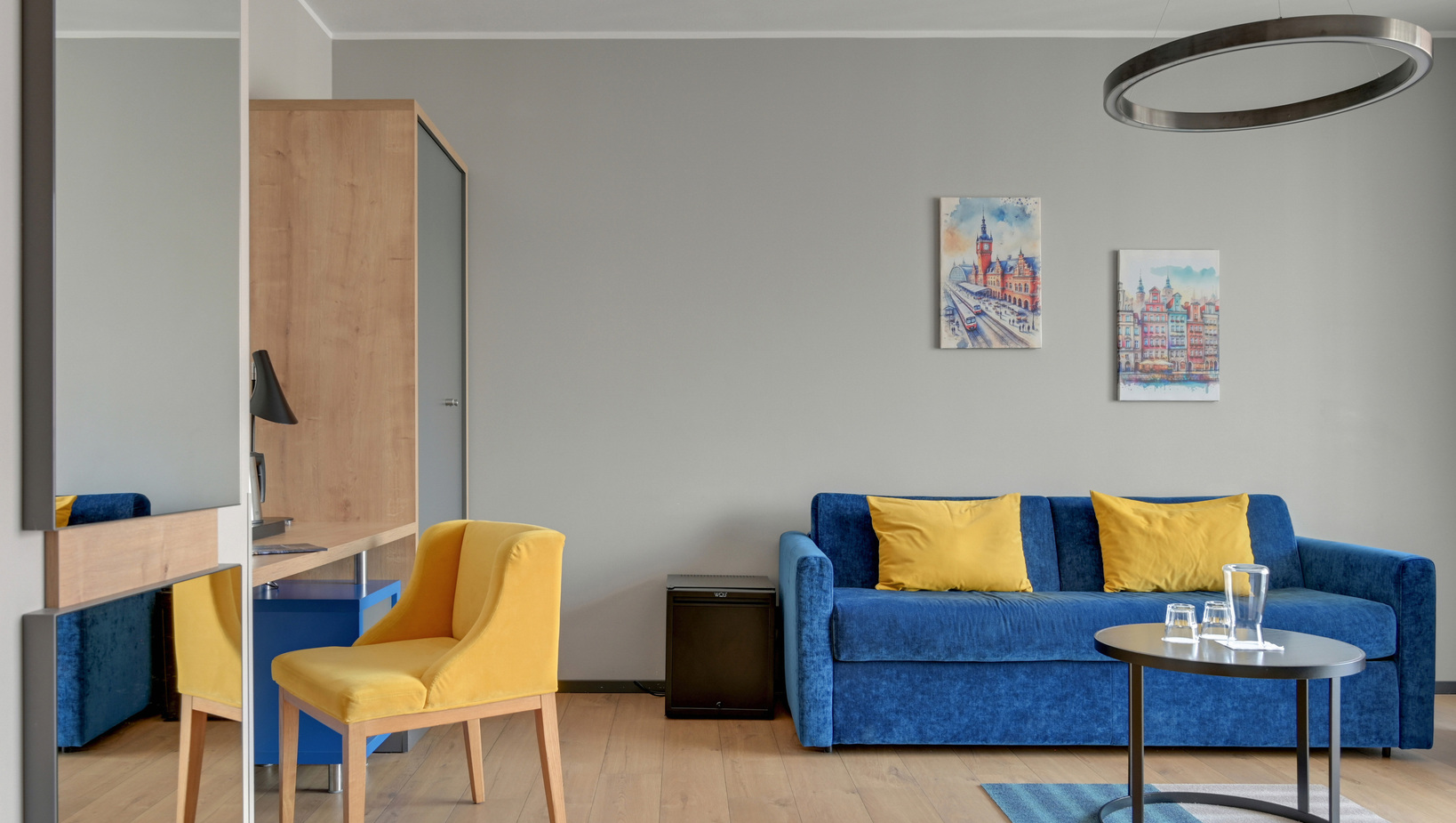 Modern hotel lounge with blue velvet sofa and yellow cushions, yellow armchair, round pendant light and a coffee table with glassware.