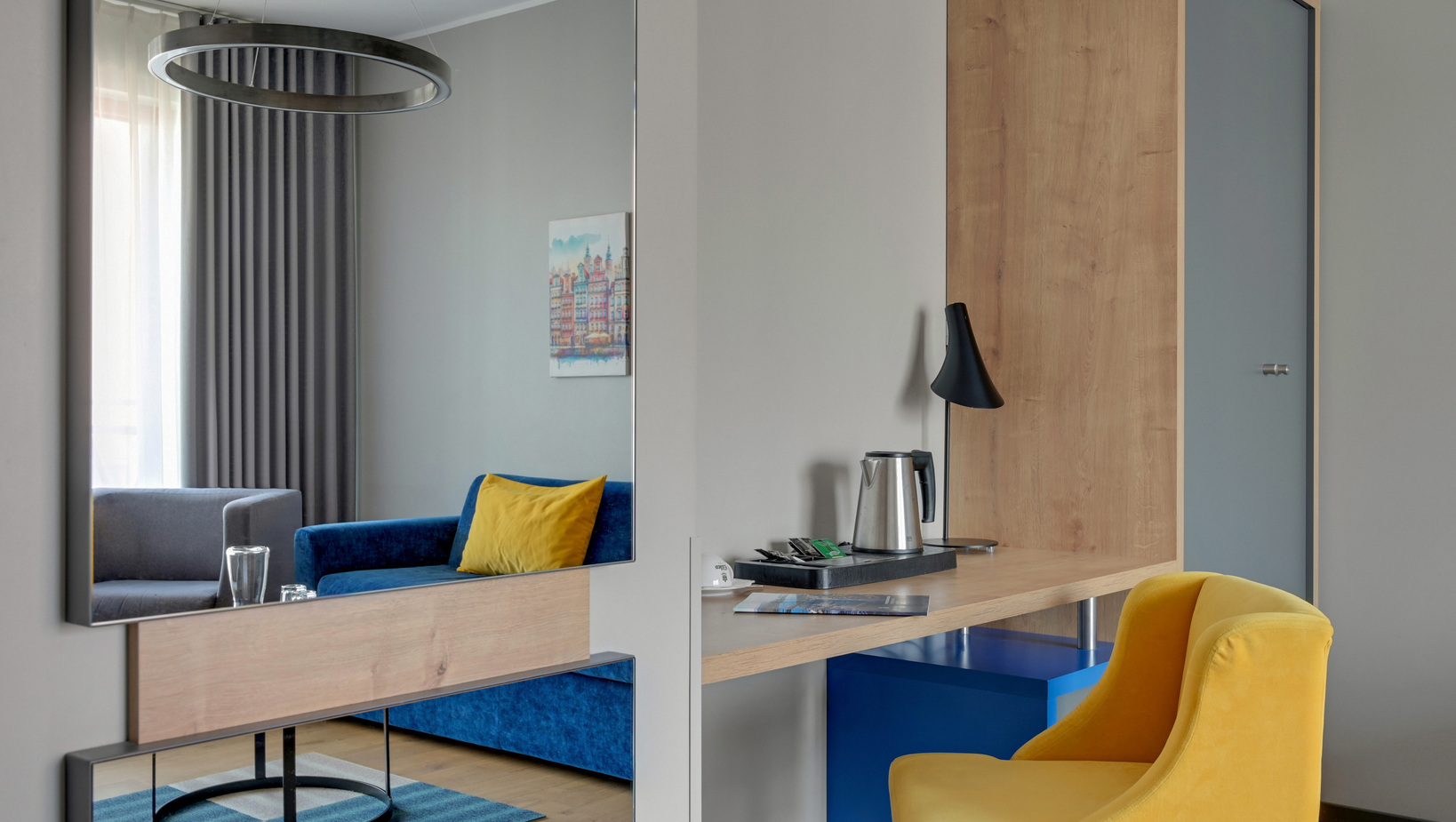 Modern hotel corner with blue velvet sofa and yellow cushion, wooden desk with electric kettle and black lamp, yellow chair.
