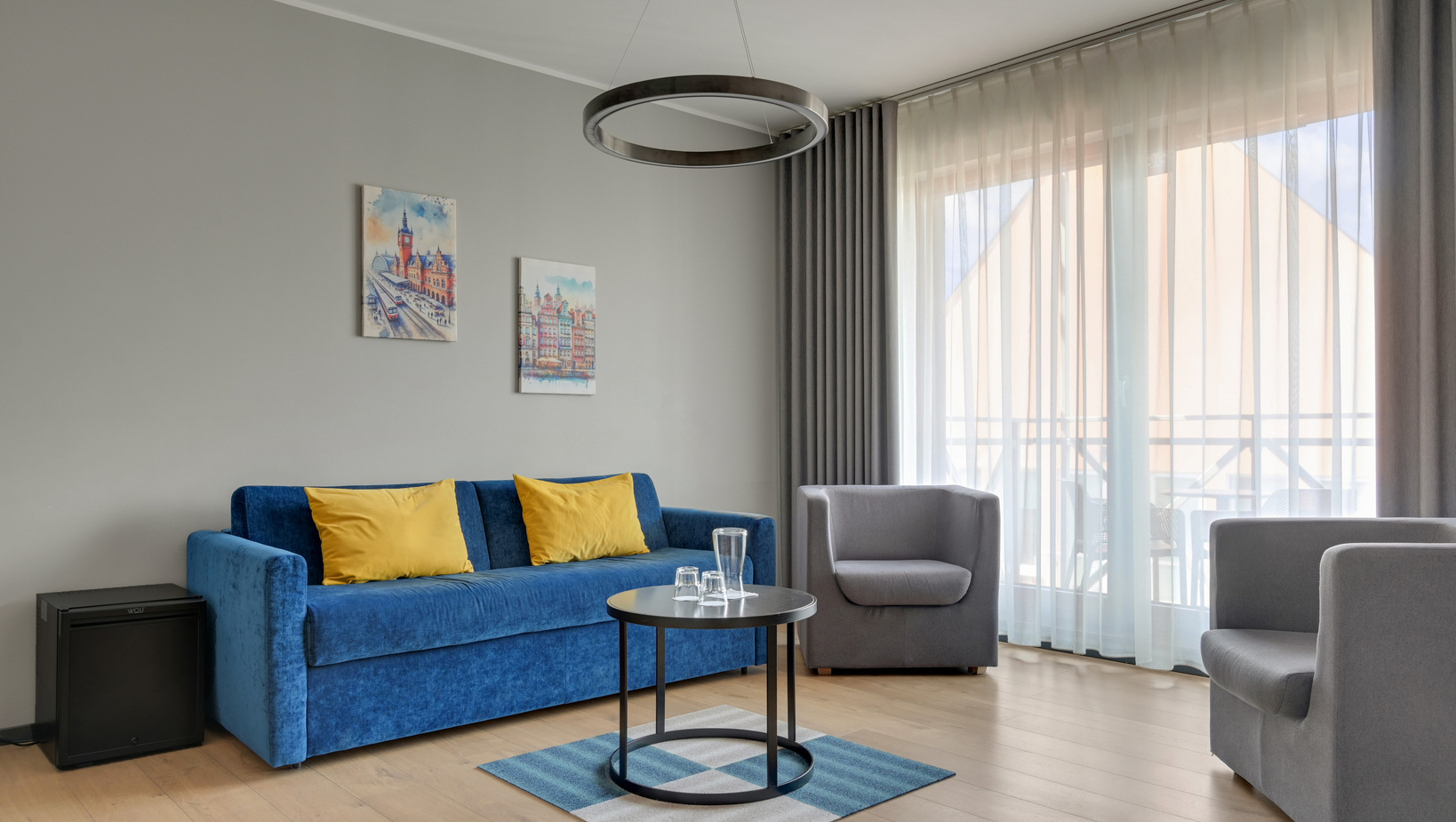 Modern hotel lounge with blue sofa and yellow cushions, gray armchairs, round pendant light, sheer curtains and balcony view.