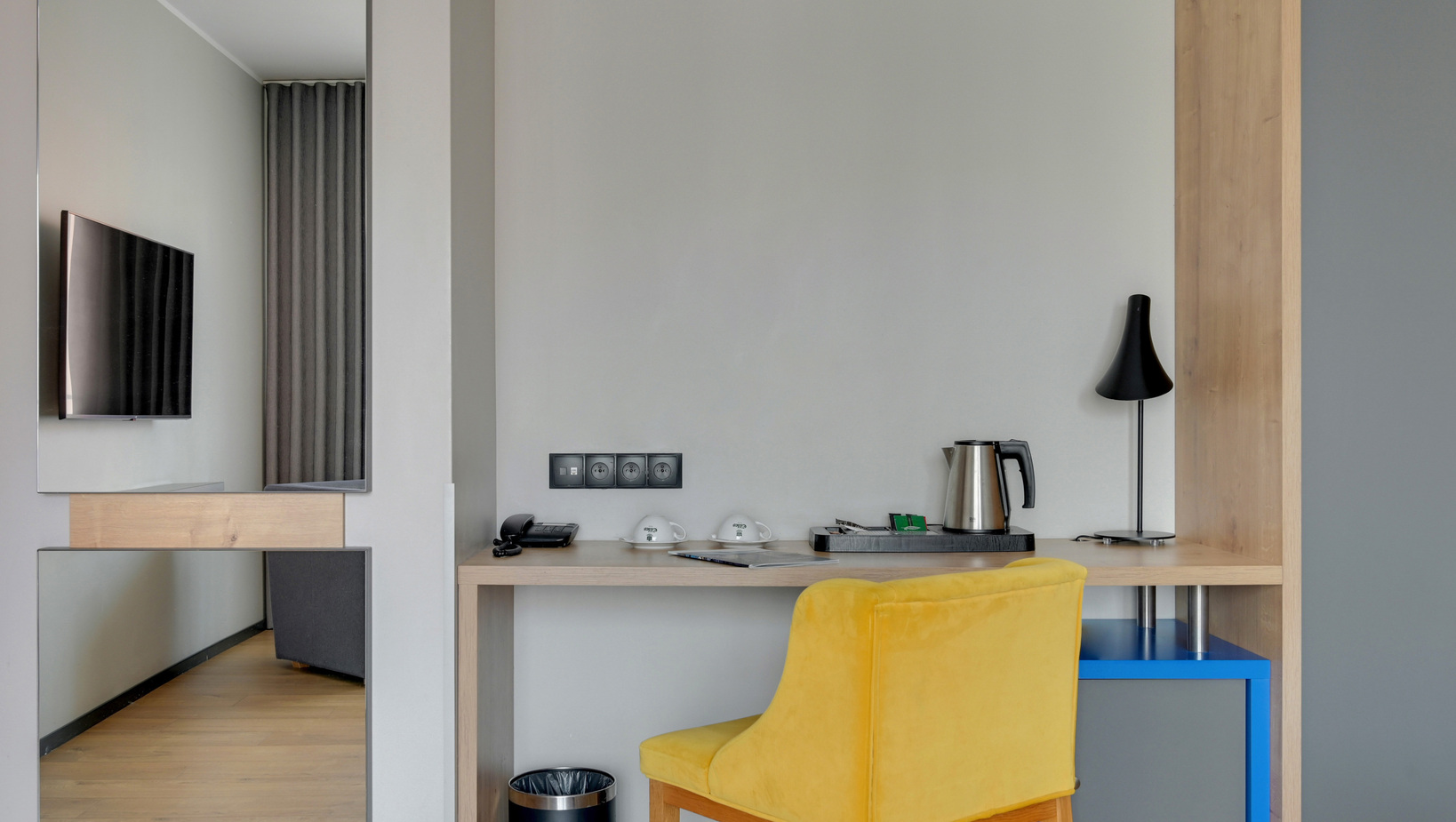 Modern hotel work desk with yellow chair, electric kettle and cups on a tray, black desk lamp, wall TV and power outlets