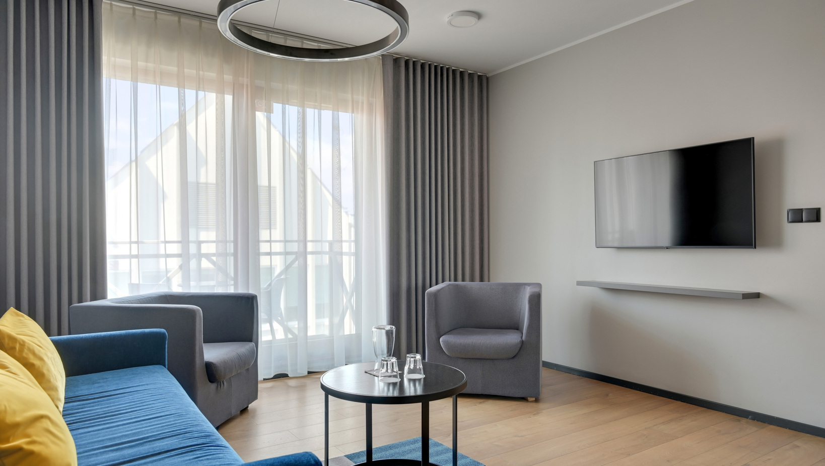 Modern hotel lounge: blue sofa, yellow cushions, gray armchairs, pendant ring light, wall TV and balcony with sheer curtains.