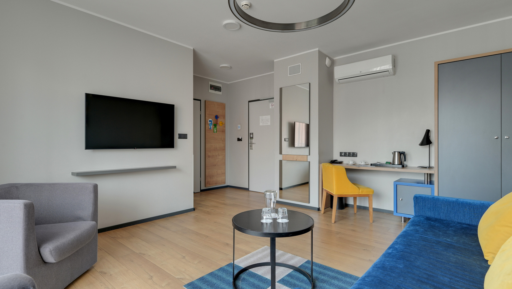 Modern hotel lounge-style room with blue sofa, yellow desk chair, wall TV, desk with kettle, wardrobe and air conditioning.