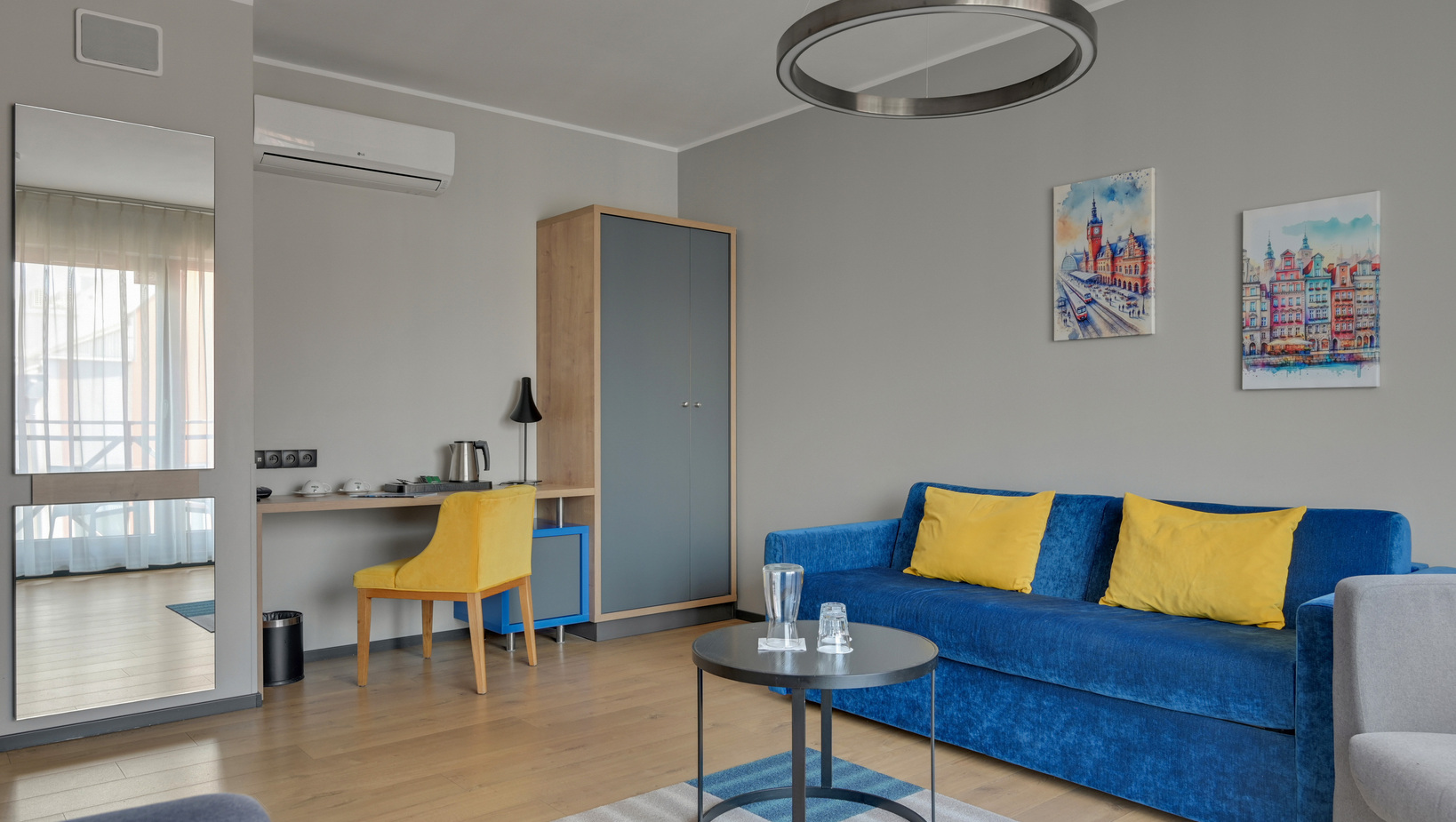 Modern hotel living area with blue sofa and yellow cushions, round pendant light, desk with yellow chair, kettle and wardrobe