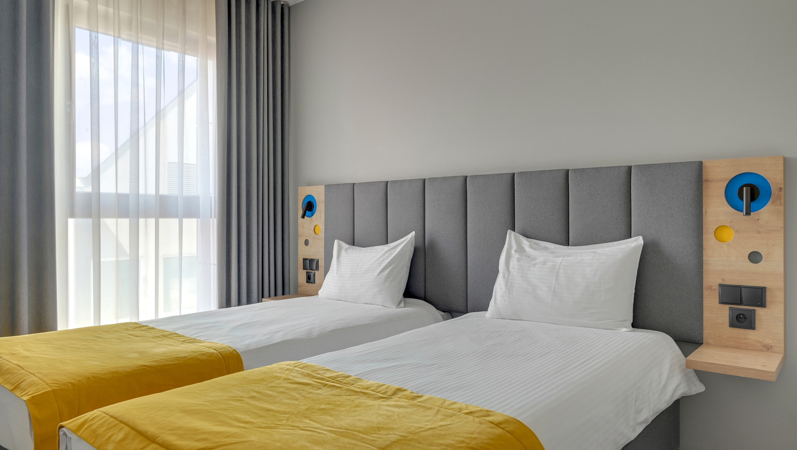 Bright twin room, grey upholstered headboard, two beds with yellow throws, bedside panels with reading lights and power outlets, sheer curtains