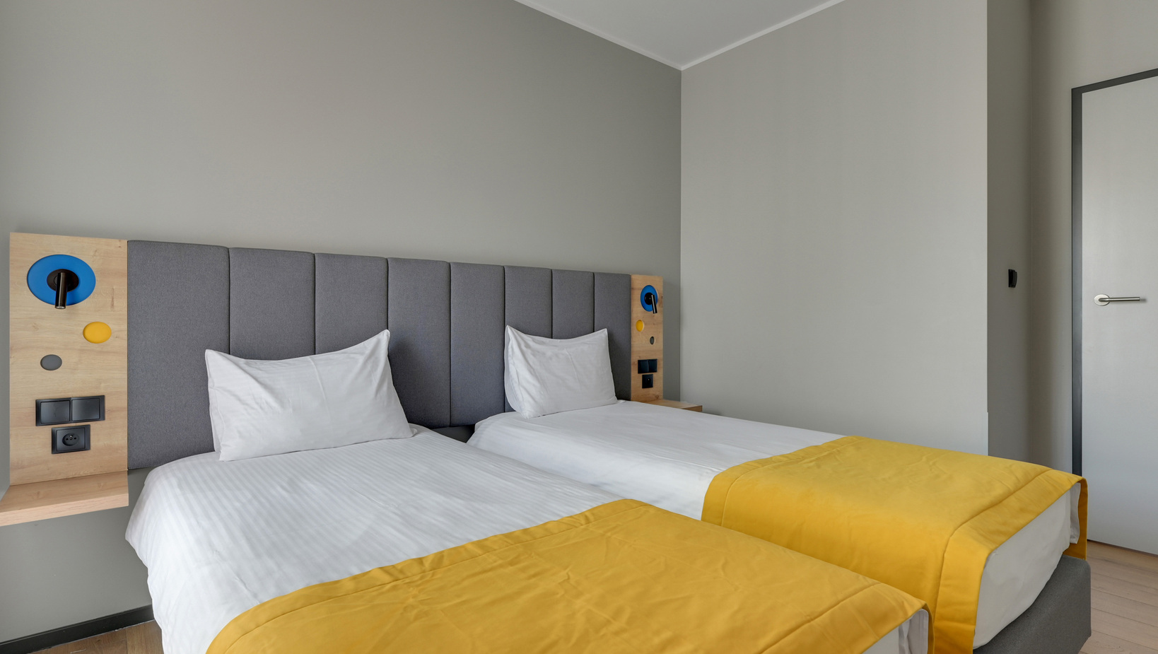 Two twin beds with grey upholstered headboard, yellow bed runners, bedside reading lamps and built-in power outlets.