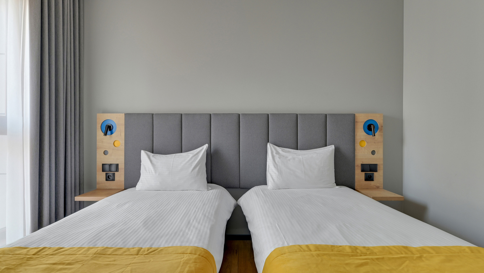 Twin beds with grey upholstered headboard, white linens and yellow throws; wooden bedside panels with blue reading lamps and outlets; soft grey curtains