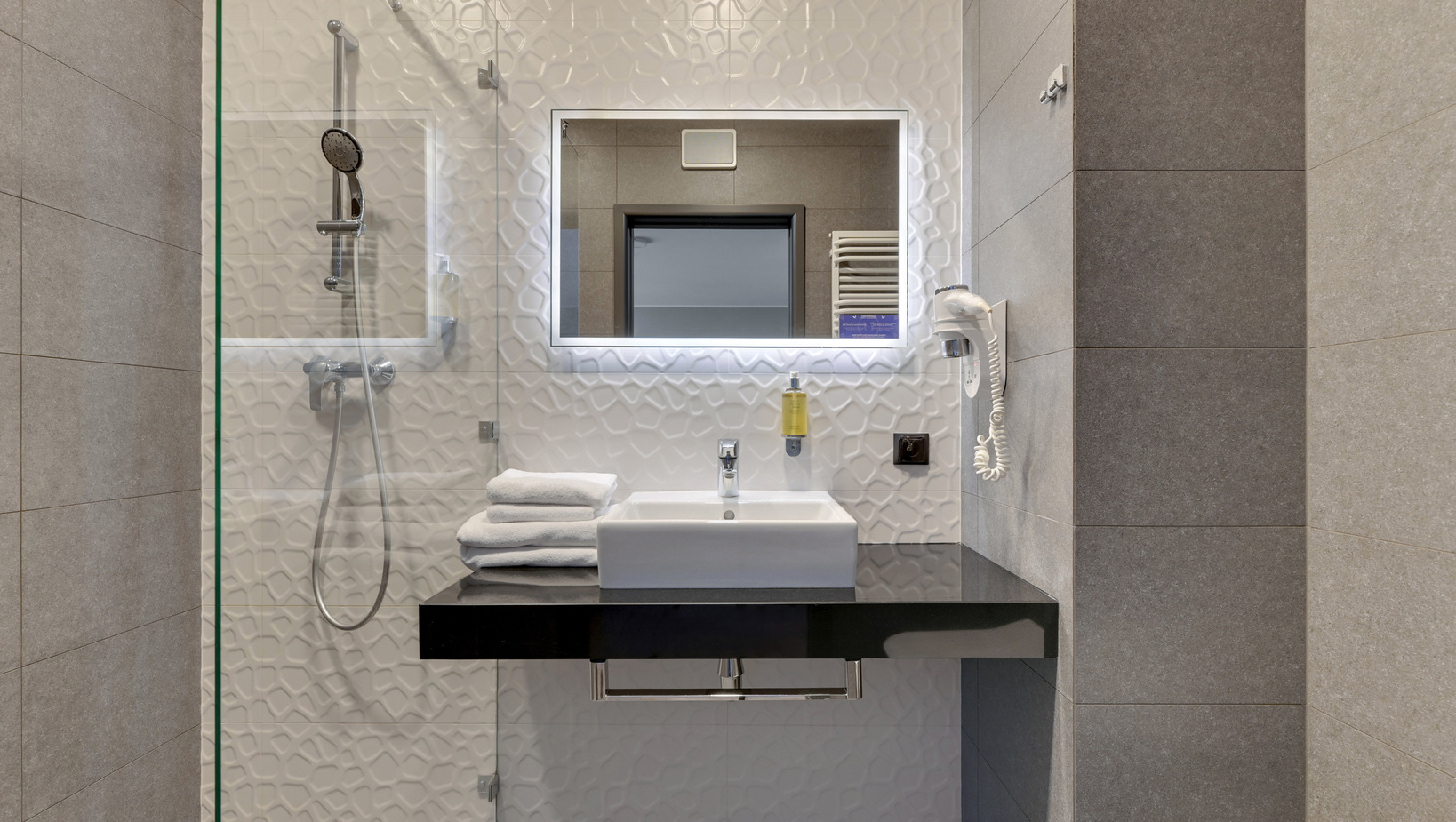 Modern hotel bathroom with glass shower, illuminated mirror, white basin on black counter, stacked towels and hairdryer.