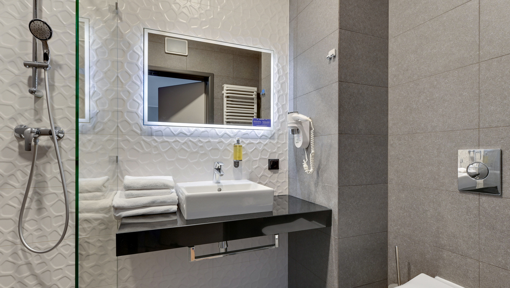 Modern hotel bathroom with walk-in shower, handheld showerhead, LED mirror, sink on black countertop, stacked towels and wall hairdryer