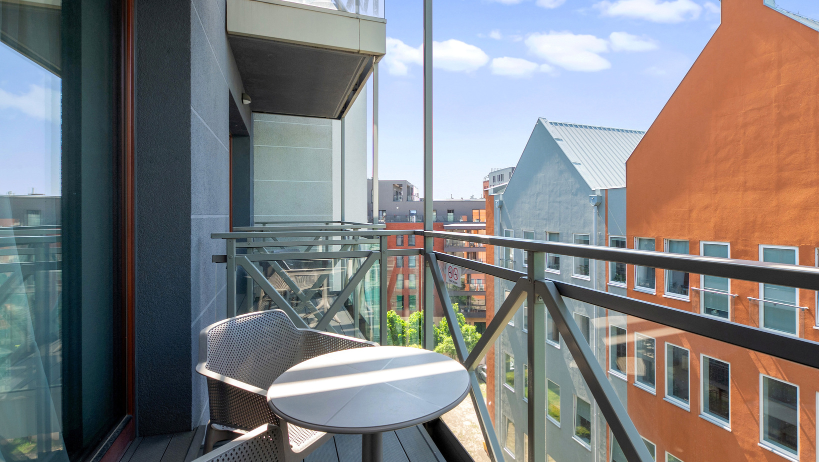 Hotel balcony with small round table and two chairs, glass-metal railing and view of colorful modern buildings
