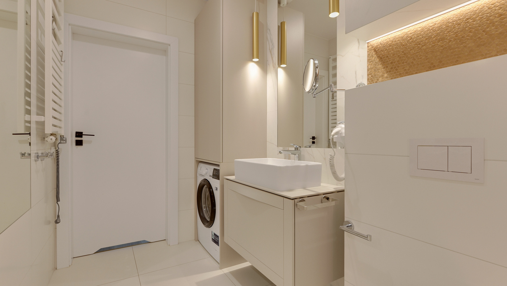 Nowoczesna łazienka hotelowa z umywalką, pralką, złotymi lampami wiszącymi, lusterkiem kosmetycznym i grzejnikiem na ręczniki.