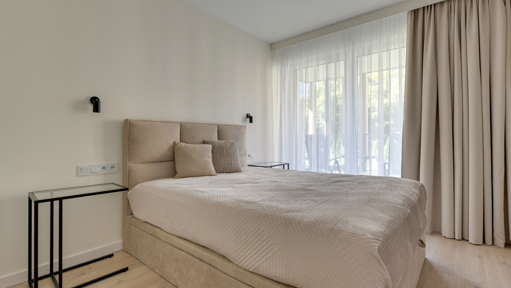Jasny nowoczesny pokój hotelowy z beżowym tapicerowanym łóżkiem, pościelą, lampkami przy łóżku i balkonem z zasłonami.