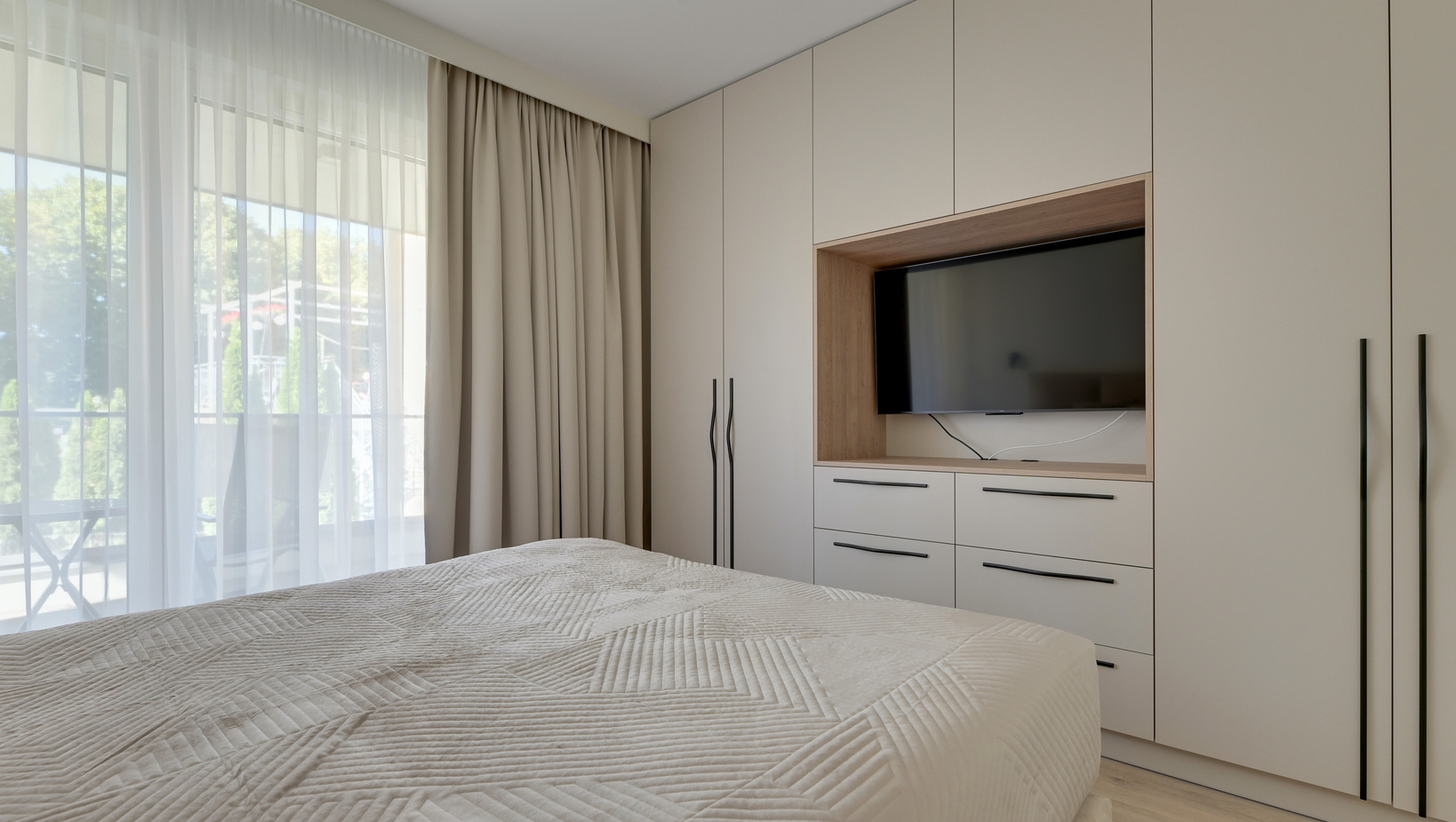 Jasny, nowoczesny pokój hotelowy z łóżkiem typu queen, zabudowaną szafą z telewizorem, firanami i widokiem na balkon.