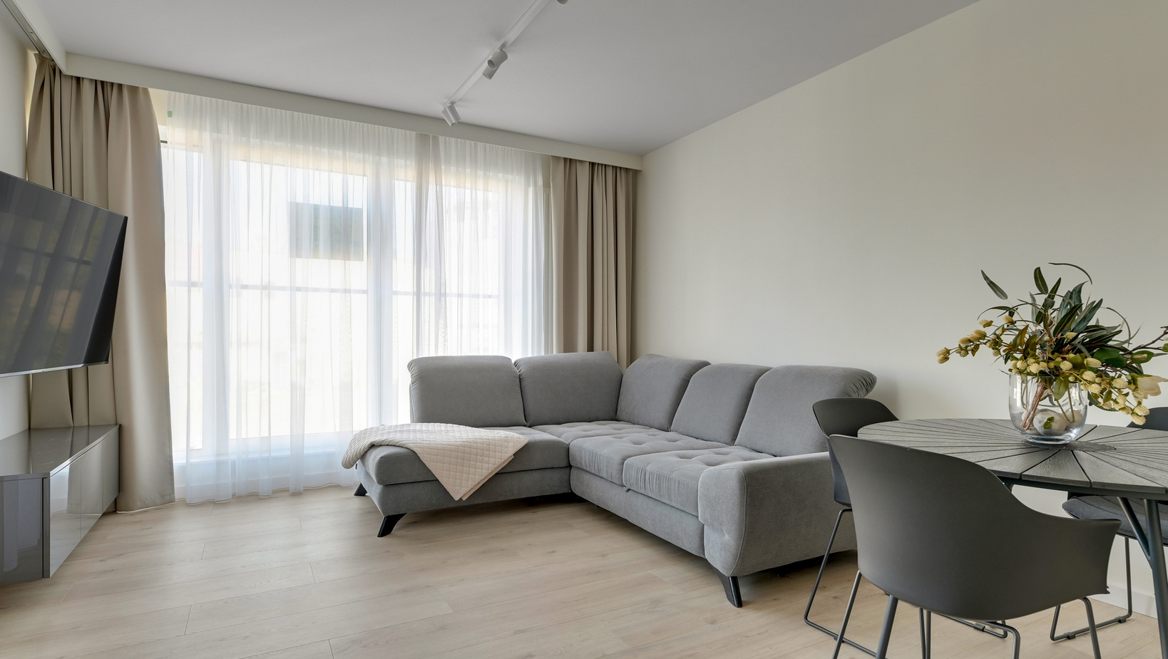 Jasny, nowoczesny salon w apartamencie z szarą narożną sofą i szezlongiem, przezroczystymi zasłonami, telewizorem i stołem z kwiatami.