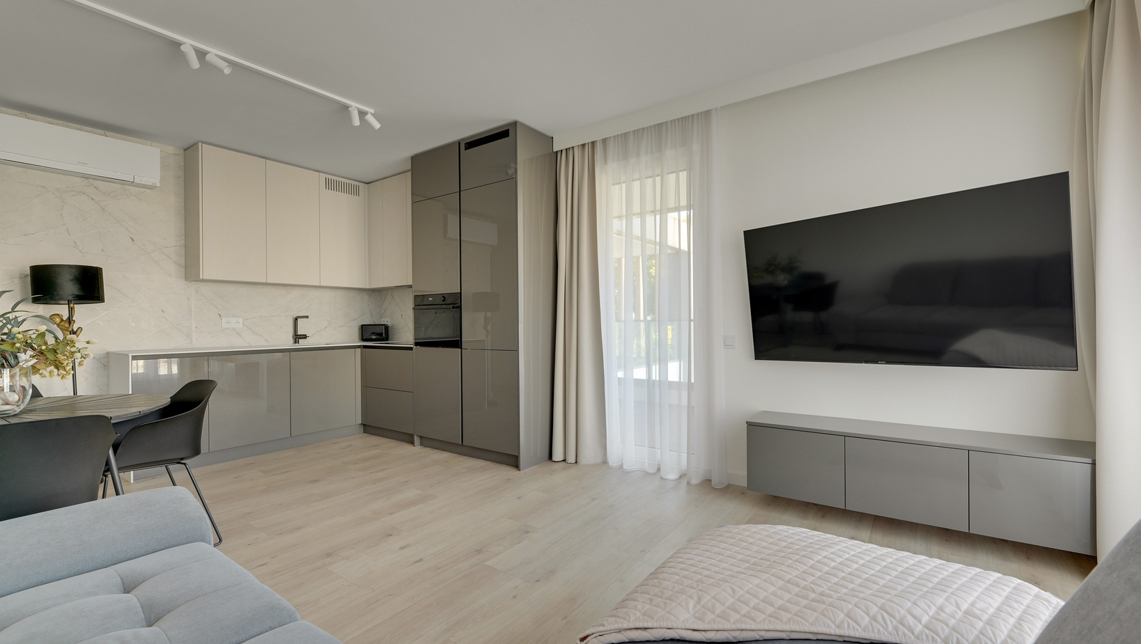 Nowoczesne studio z aneksem kuchennym, błyszczącymi szafkami, telewizorem na ścianie, stołem jadalnym, sofą i lekkimi zasłonami na balkon.