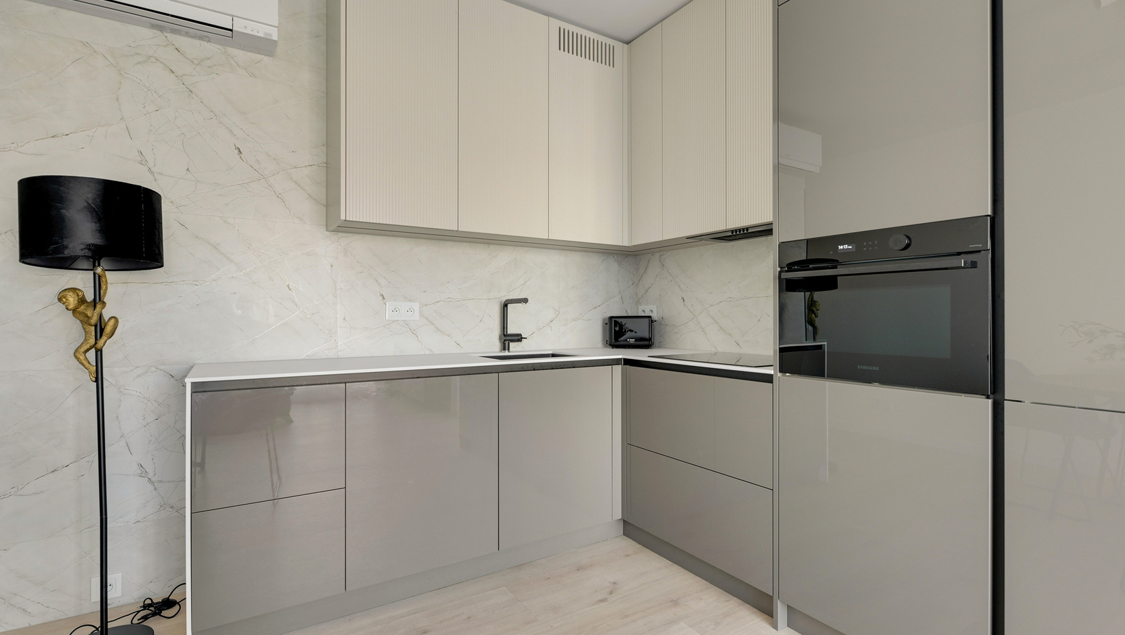 Kompaktowa nowoczesna kuchnia w apartamencie z połyskującymi szarymi szafkami, marmurowym panelem, piekarnikiem, zlewem i czarną baterią