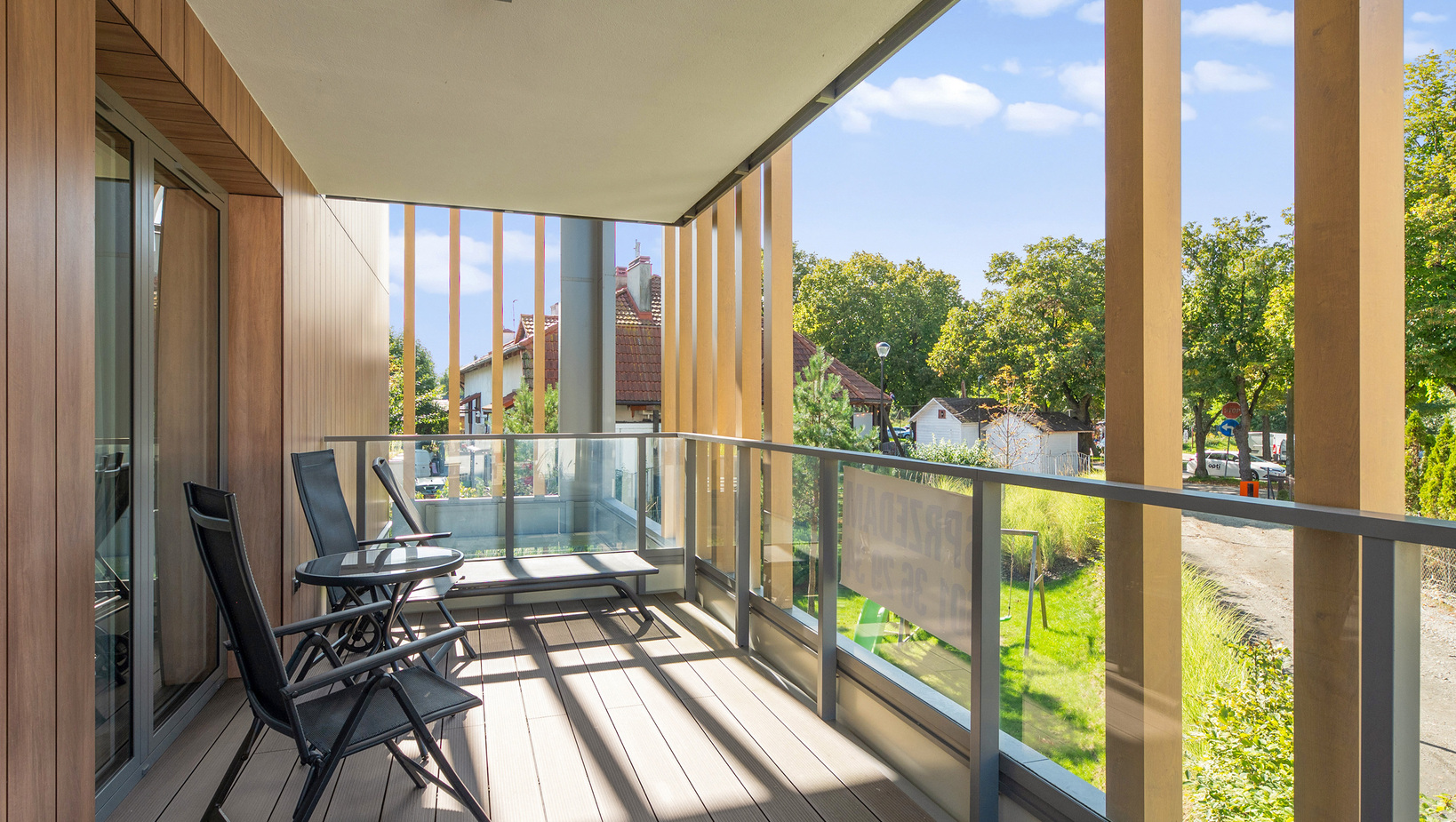 Zadaszony balkon z dwoma fotelami, stolikiem, szklaną balustradą i pionowymi drewnianymi słupami, widok na drzewa i domy.