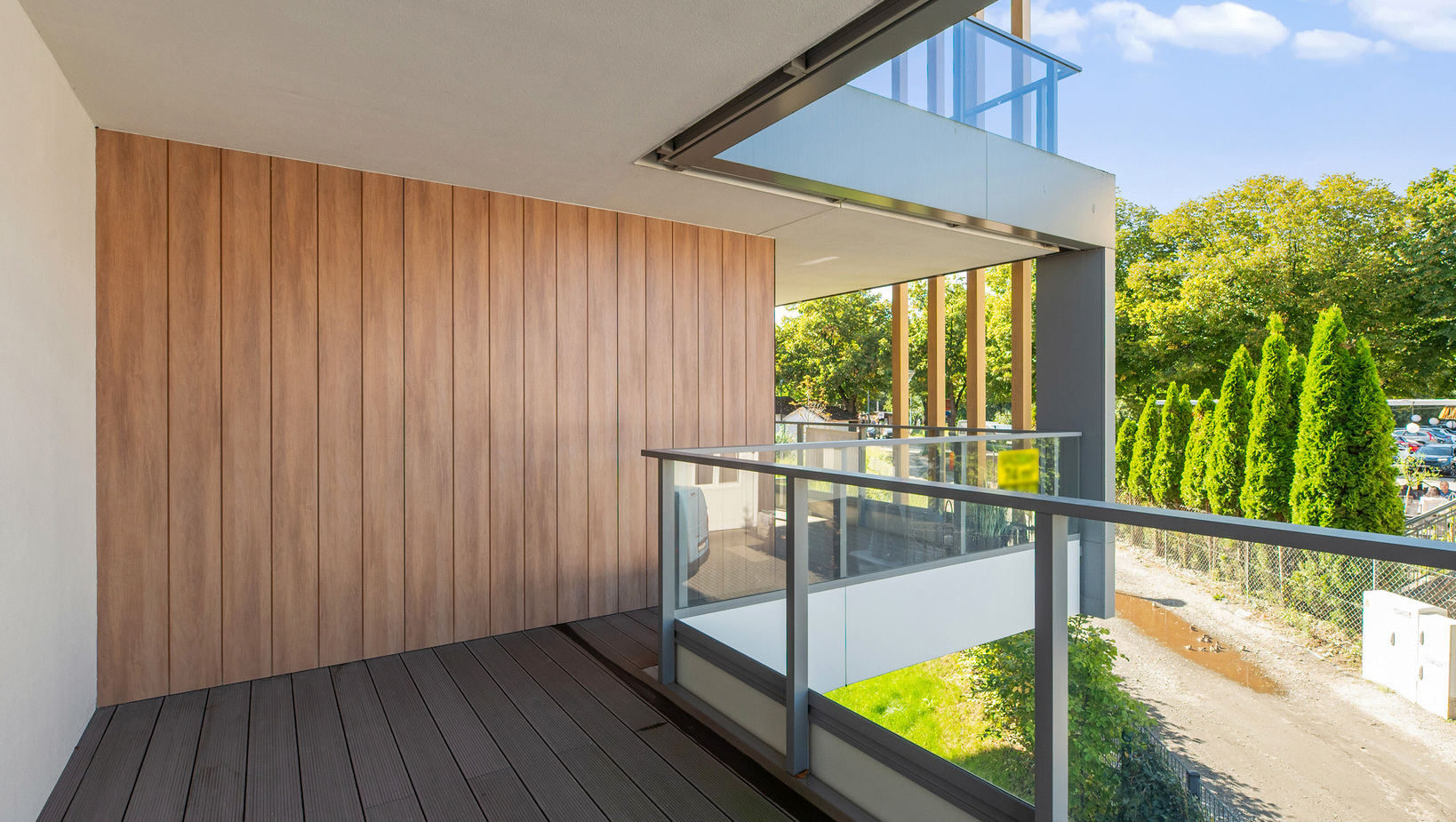 Prywatny balkon z drewnianym tarasem i pionową boazerią, przeszkloną balustradą i widokiem na korony drzew